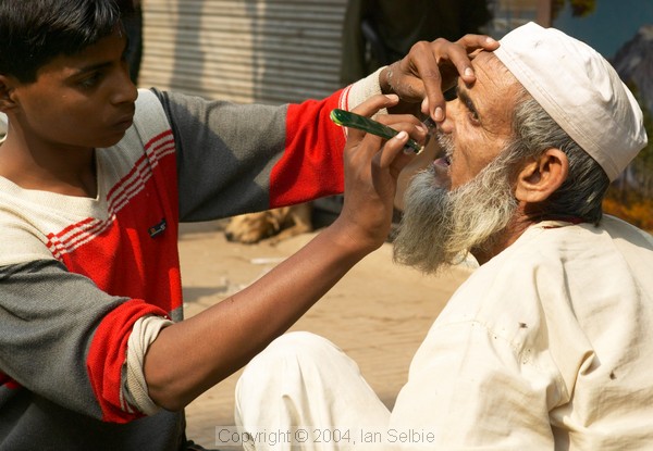 Young boy shaving the moustache of an old bearded man on the footpath by the street, Old Delhi